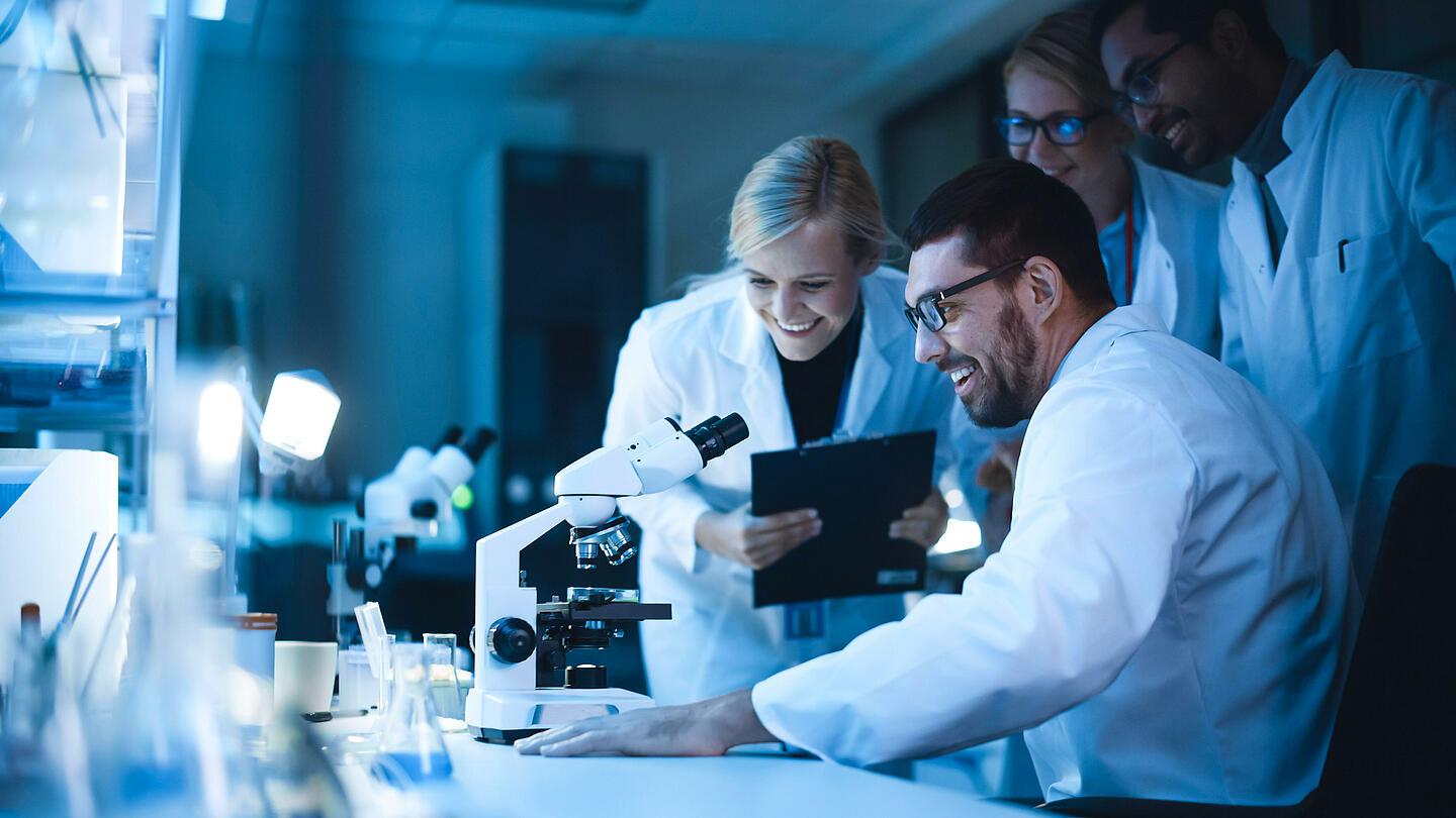 Cuatro personas sonrientes detrás de un microscopio en un laboratorio