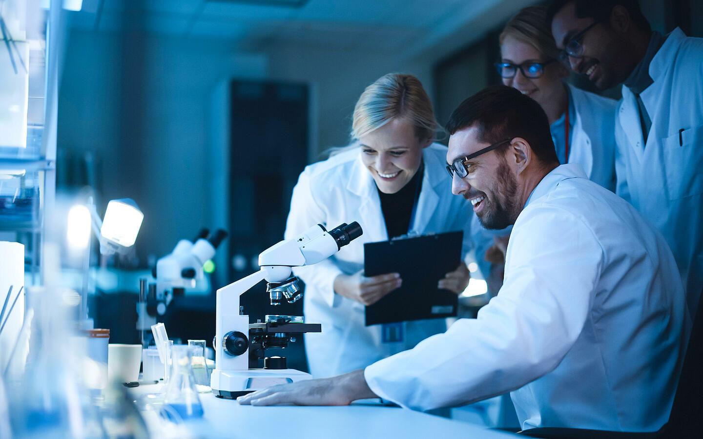 Cuatro personas sonrientes detrás de un microscopio en un laboratorio