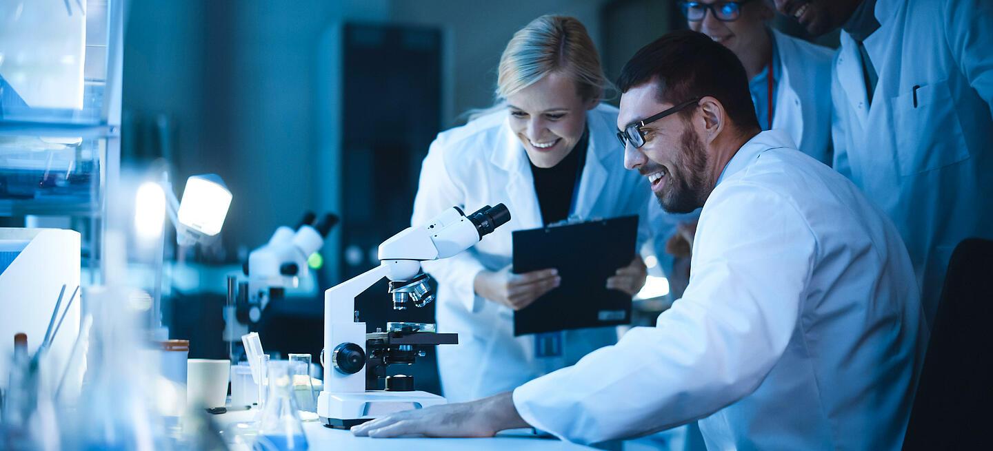 Cuatro personas sonrientes detrás de un microscopio en un laboratorio