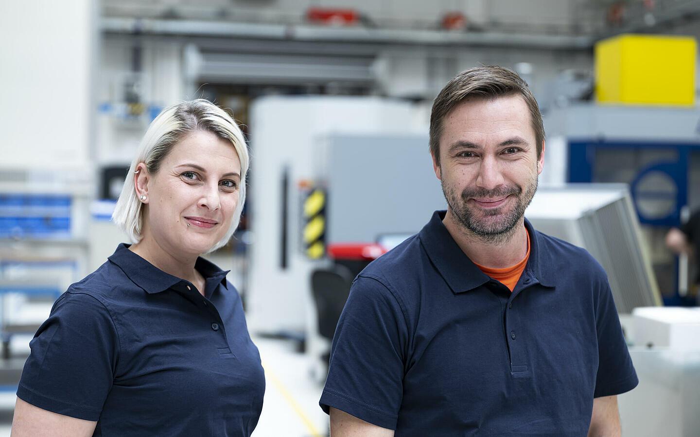 Zwei Mitarbeiter, die blaue Polo-Shirts tragen, stehen nebeneinander und lächeln in die Kamera